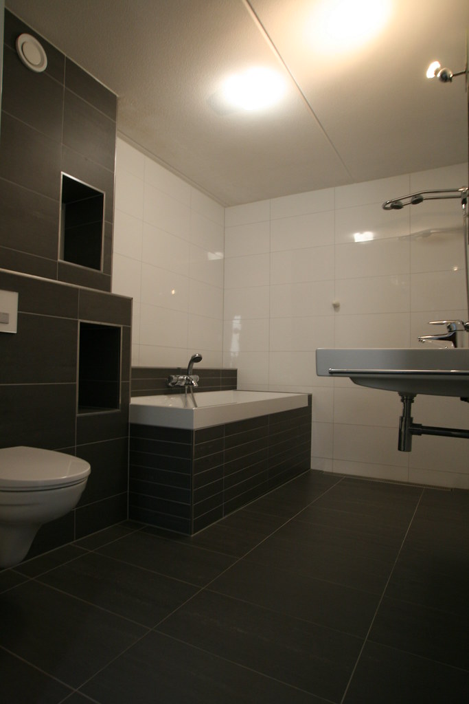 Badkamer met donkere tegels, houtlook vloer, ligbad en hangtoilet